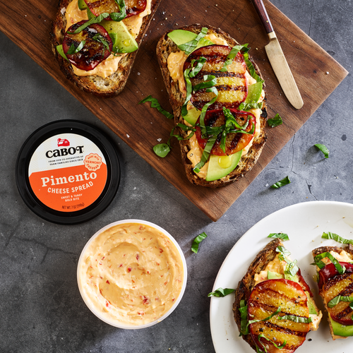 A rustic wooden board displays open-faced sandwiches with grilled tomatoes, avocado, and fresh greens. A tub of Cabot Creamerys Pimento Cheese Spread is on the left, alongside a bowl of creamy Vermont Cheddar. A knife is nearby for easy serving.